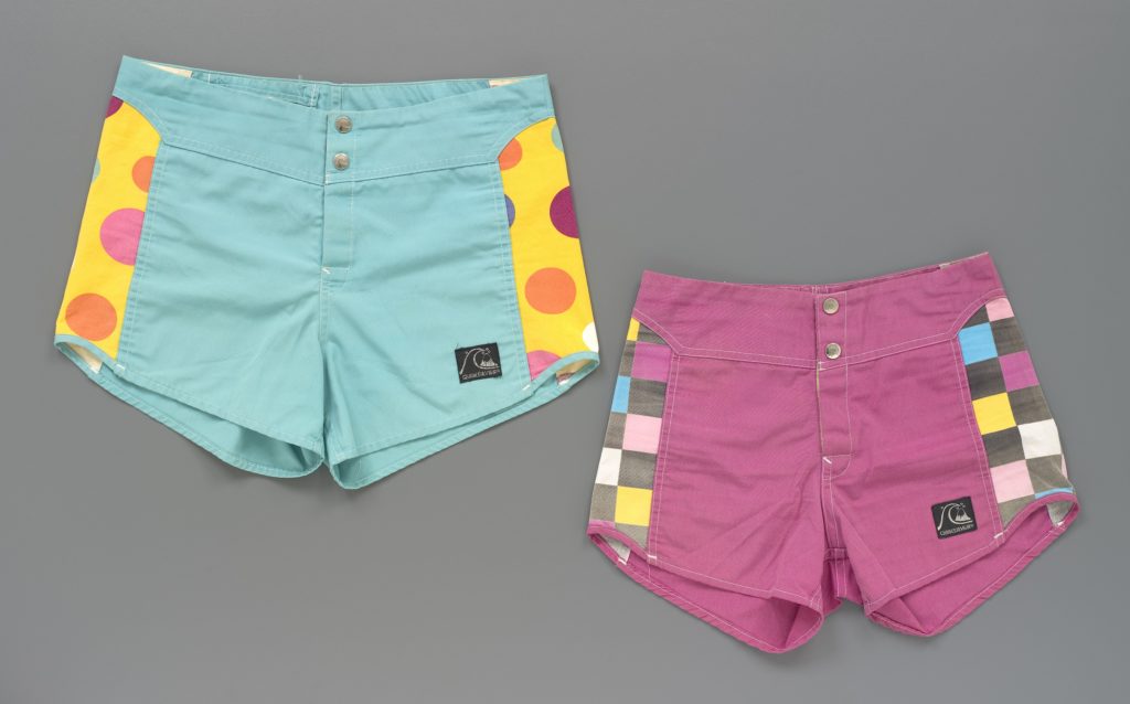 Two pairs of men's board shorts in green and pink with inserts on the sides consisting of multi-coloured polka dots or squares. There are two press-stud fastening and a Velcro fly at the centre front. A black brand label is stitched to the front left leg displaying a logo and the text 'Quiksilver' in silver thread.